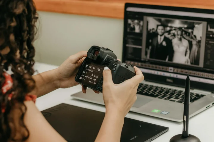 Woman holds DSLR camera at desk with MacBook showing black-and-white wedding photo and Wacom tablet nearby.