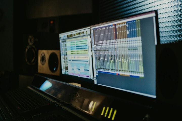 Dual monitors show audio tracks and mixer in a dimly lit music studio.