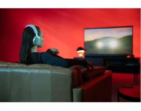 Side view of woman with long hair wearing white headphones, sits comfortably on couch facing TV screen with hills and sunset glow, red ambient lighting.