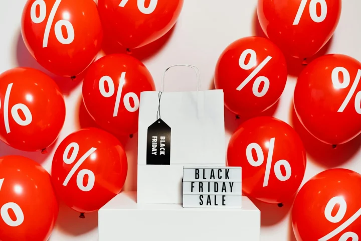 Red balloons with percent signs float around a Black Friday sale sign and white shopping bag.