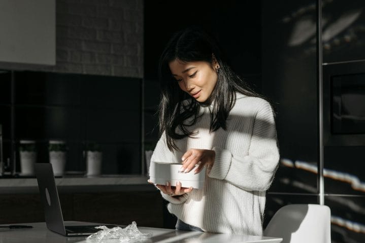 Woman in cream sweater smiles while holding white mug in modern kitchen.