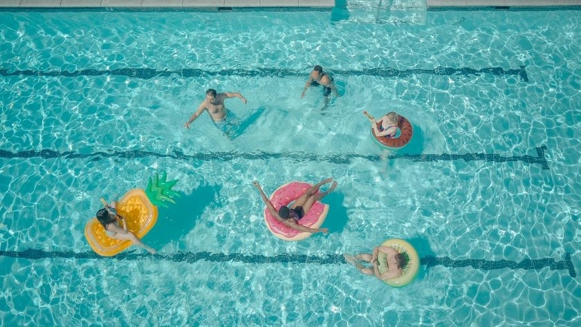 Aerial view of people floating in colorful inflatables like pineapples and flamingos in a clear blue pool.