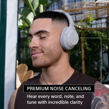 Smiling man listens intently through white Sony headphones in outdoor plant setting with tattoo visible.