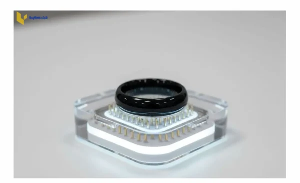 A black Samsung Galaxy Ring charging inside its innovative transparent charging case with a soft white LED indicator light on a white desk.